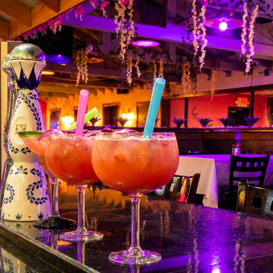 Two vibrant red cocktails with colorful straws on a bar, with purple lighting and restaurant interior.