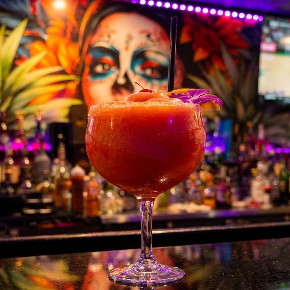 A vibrant red frozen drink with a cherry and flower garnish sits on a bar with colorful decor.