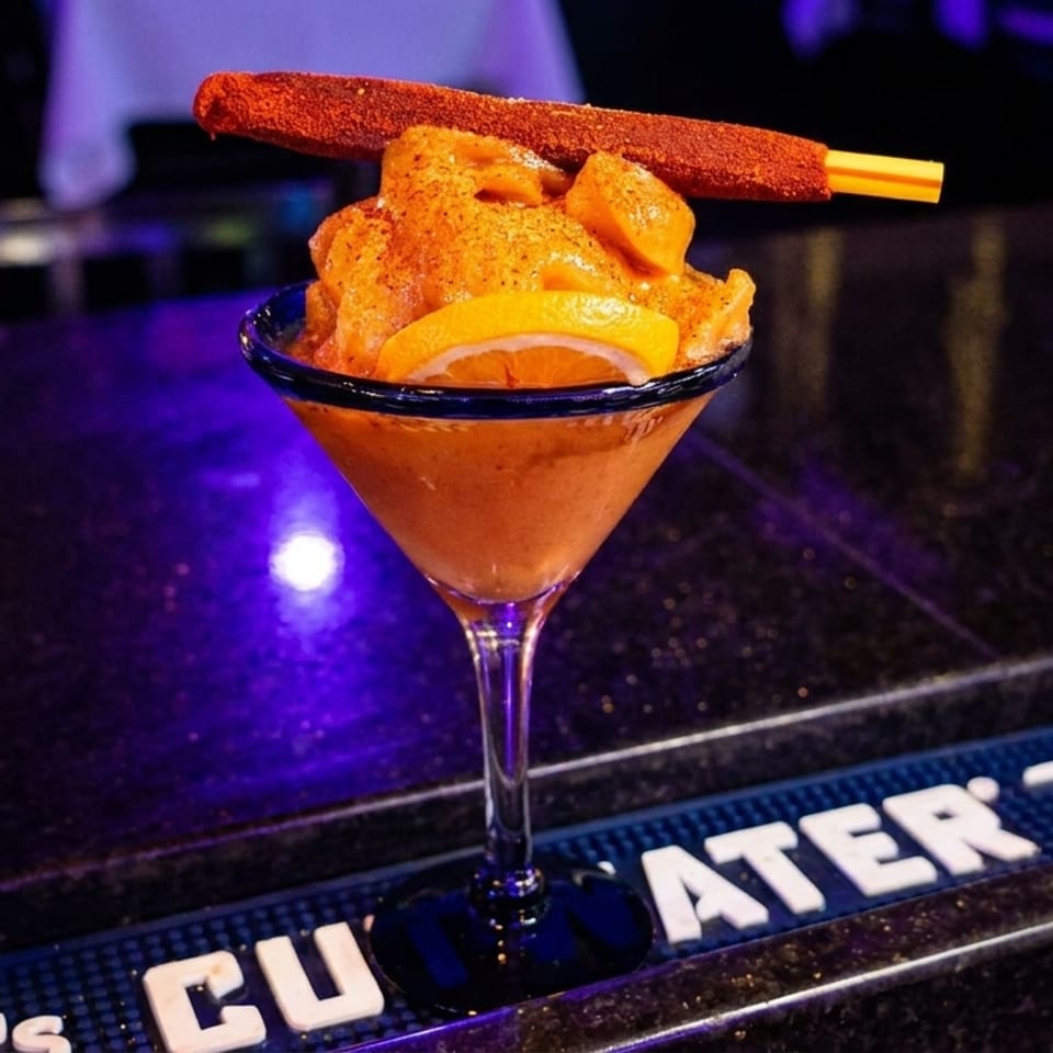 A vibrant orange frozen drink in a martini glass, garnished with an orange slice and a chili-dusted stick.