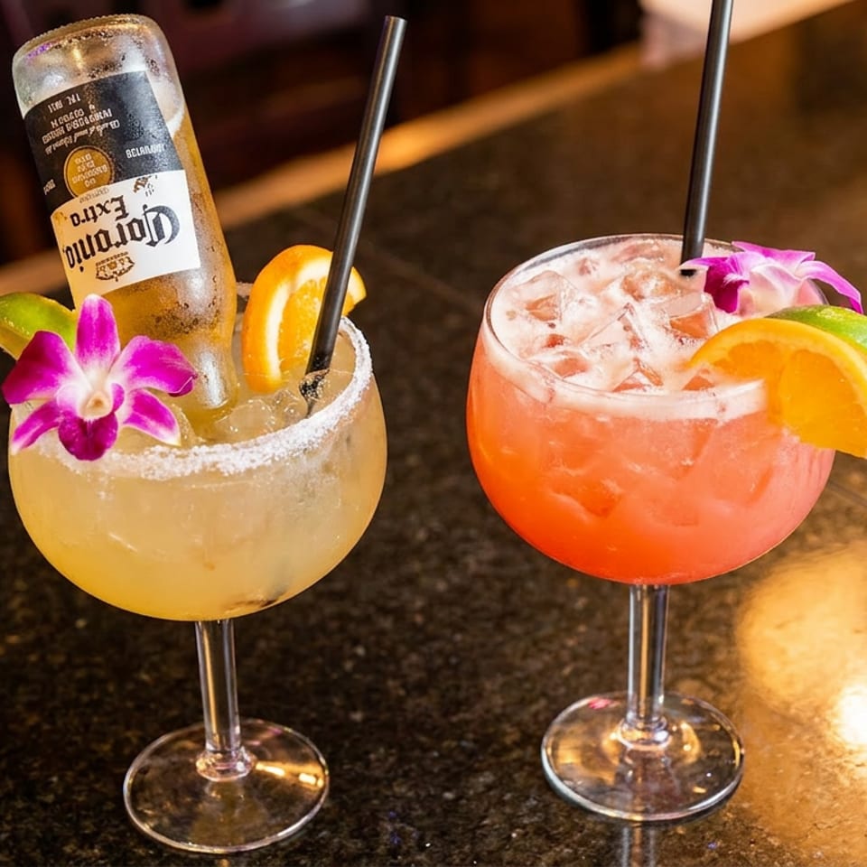 Two colorful cocktails, one with a Corona bottle, garnished with fruit and orchids on a dark bar.