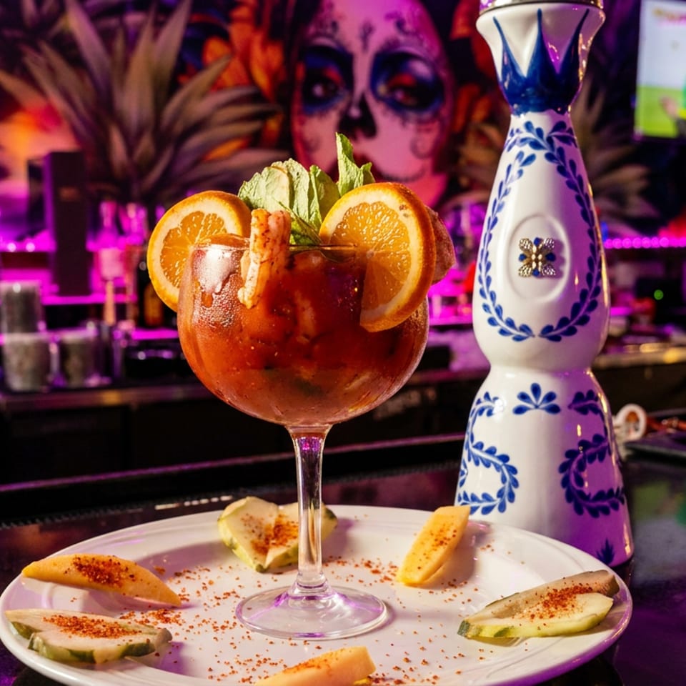 A vibrant red cocktail garnished with orange slices and shrimp, served with spiced fruit on a bar.