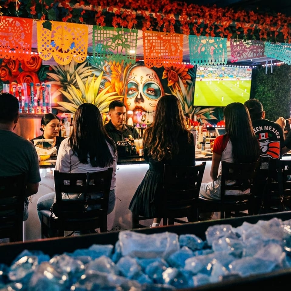 Patrons seated at a lively bar with colorful papel picado decorations and a large mural.
